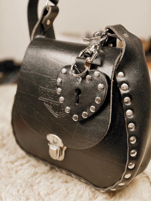 a black studded leather key chain in the shape of a heart is attached to a studded heavy leather harly davidson bag. 